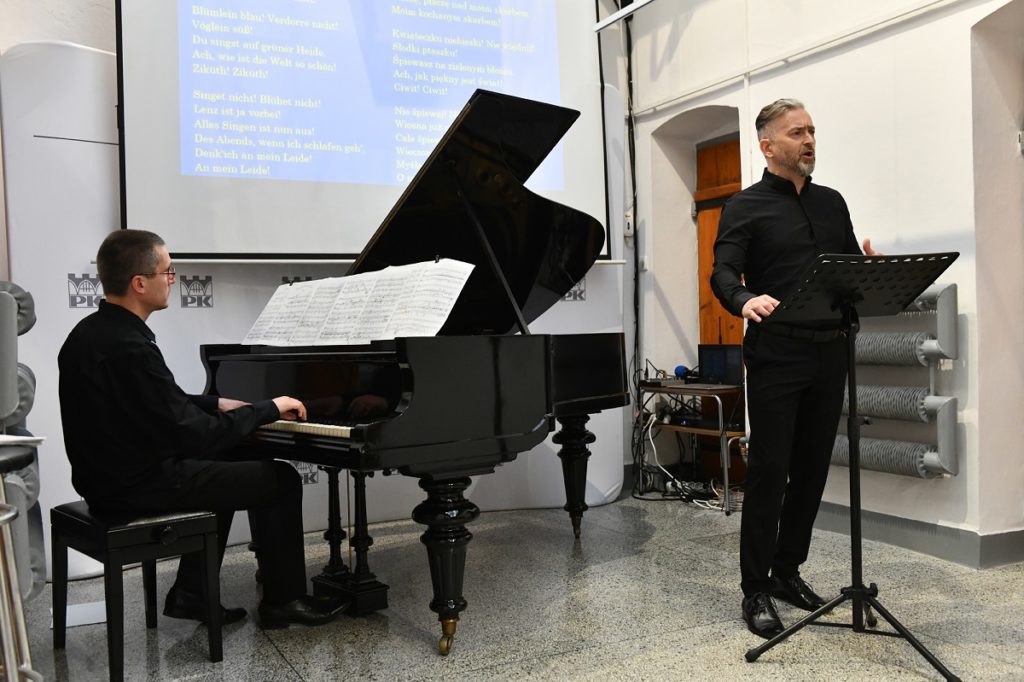 Koncert. Jeden z mężczyzn gra na fortepianie, a drugi śpiewa do mikrofonu